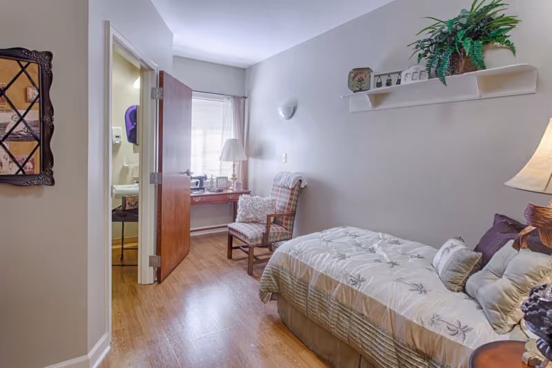 Cozy single bedroom with a bed, upholstered chair, desk by a window, and an open door leading to a bathroom.