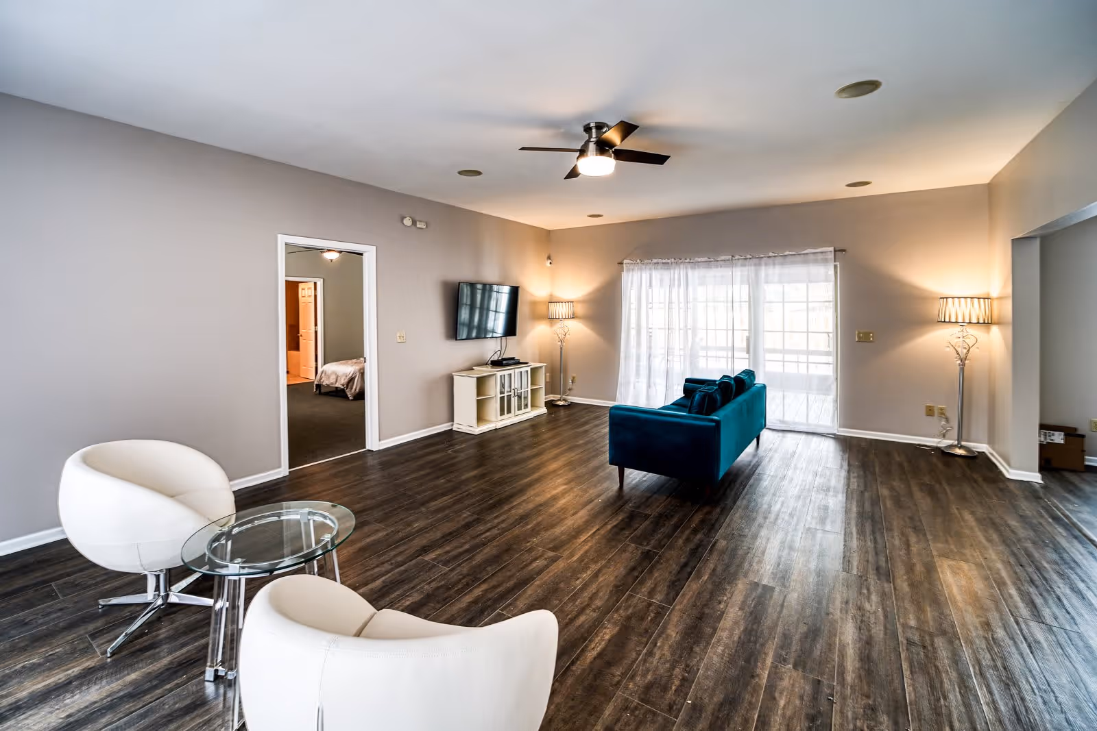A spacious living room with dark wood flooring, a teal sofa facing a wall-mounted flat-screen TV, two white modern chairs around a small glass table, a ceiling fan with light, two floor lamps, and large windows with sheer white curtains letting in natural light. A doorway leads to a bedroom with a visible bed.