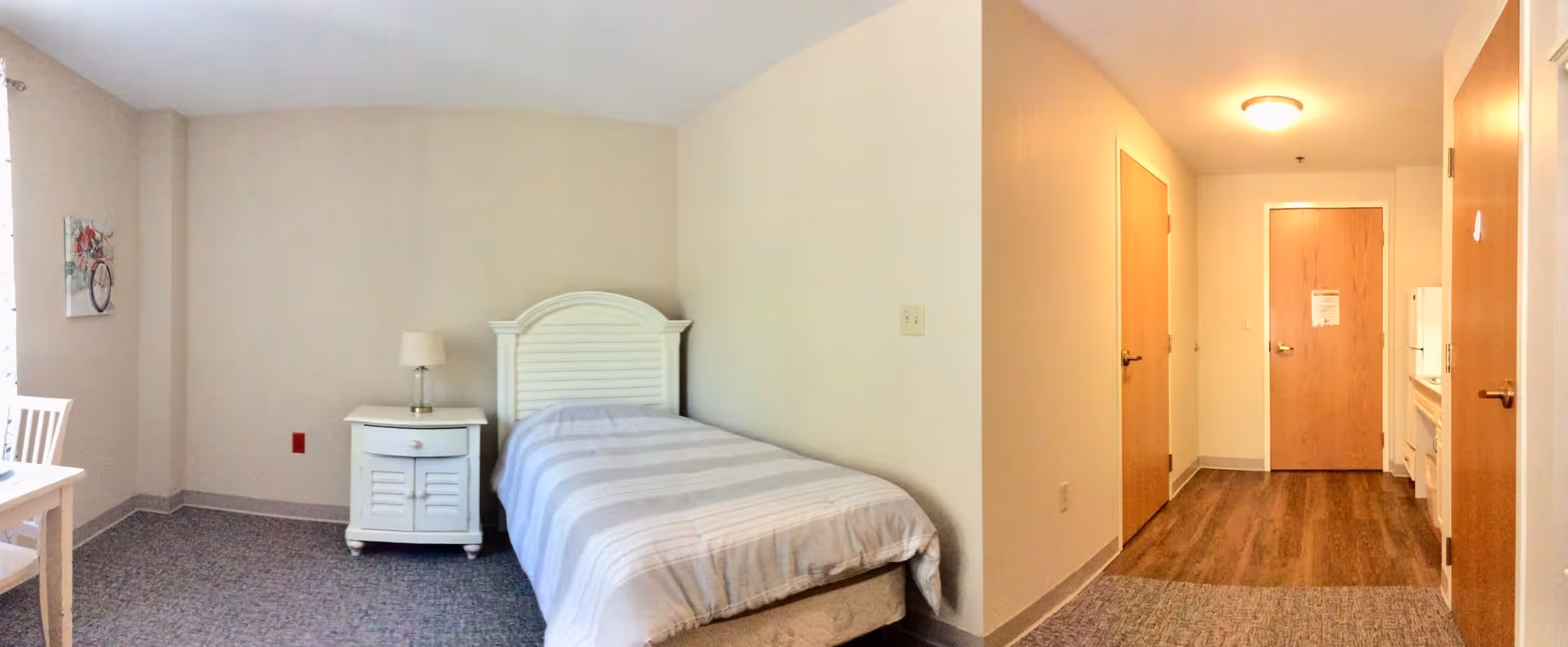 A small, simple bedroom in an assisted living facility with a single bed featuring a white headboard and striped bedding. Next to the bed is a white nightstand with a lamp. The room has beige walls, a piece of wall art with a bicycle and flowers, and a small dining table with chairs. The floor is carpeted near the bed and transitions to wood flooring near the entrance door and kitchenette area.