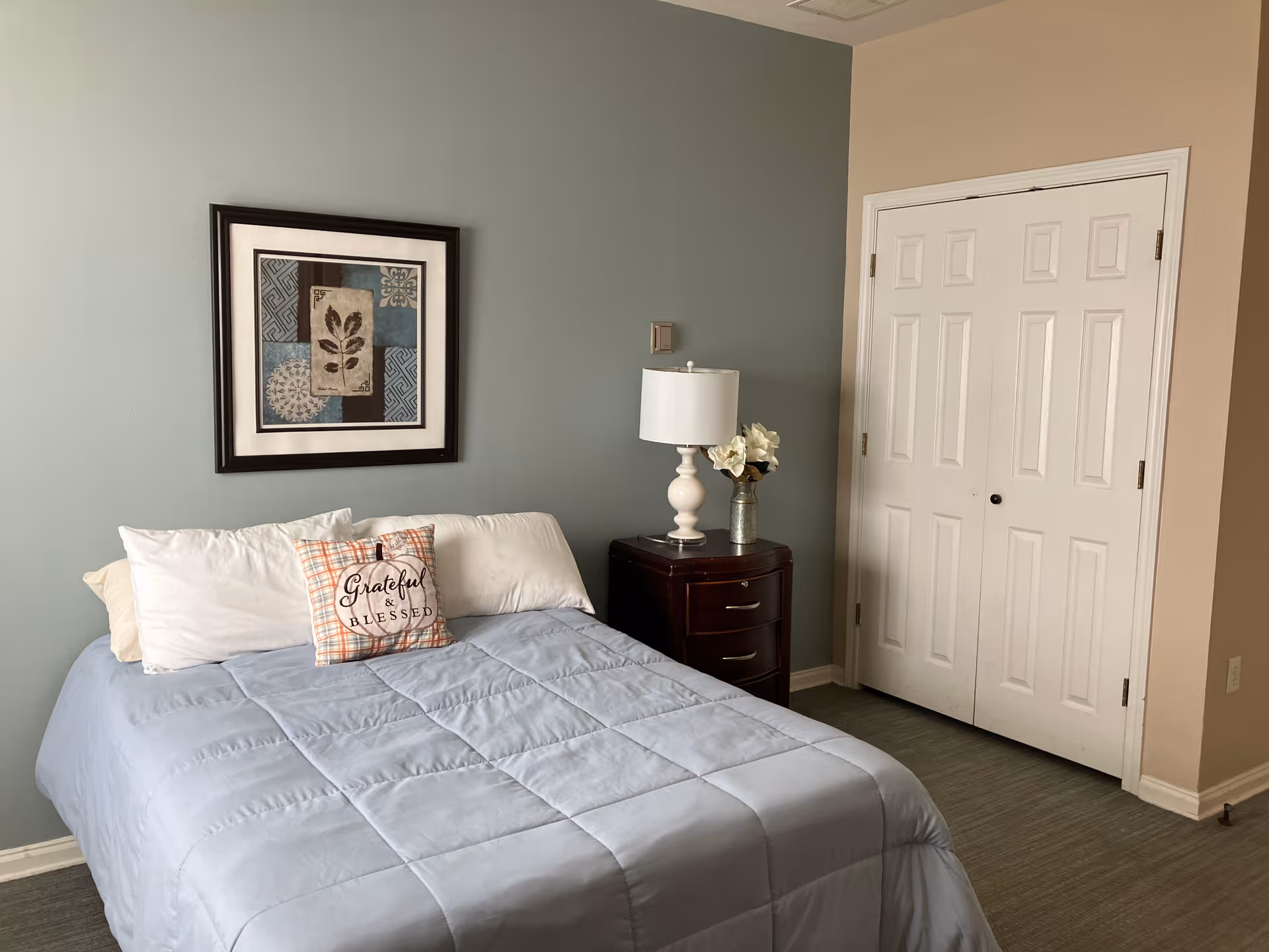 A neatly made bed with a light blue comforter, white pillows, and a decorative pillow that says 'Grateful & Blessed' in a bedroom. There is a dark wooden nightstand with a white lamp and a small vase with flowers next to the bed. A framed abstract artwork hangs on the light blue wall above the bed. A white double-door closet is visible on the right side of the room.