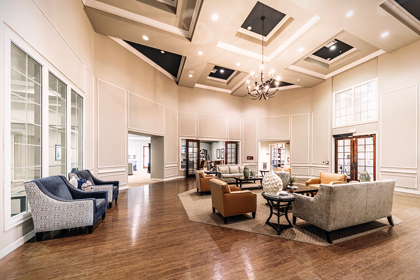 Spacious and well-lit living room area with high ceilings and recessed lighting. The room features a chandelier, multiple seating options including sofas and armchairs arranged around coffee tables on area rugs. Large windows and glass doors allow natural light to enter the room. The walls are decorated with molding details, and the floor is wooden.