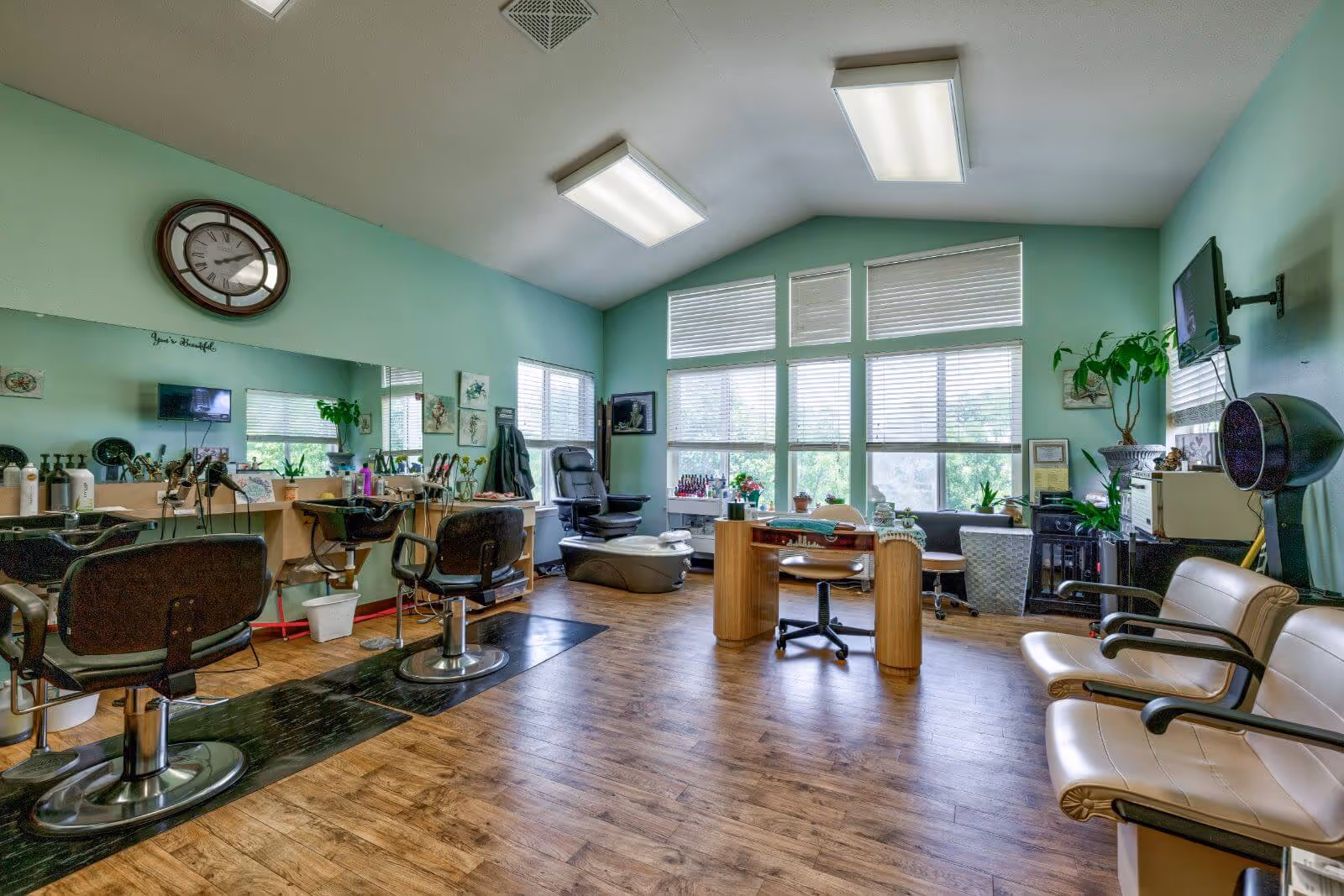 A bright and spacious salon room with light green walls and large windows with blinds. The room features two black salon chairs in front of a long mirror with hair care products and tools on the counter. There is a pedicure chair near the windows, a wooden manicure table with a rolling chair, and two beige waiting chairs on the right side. The floor is wooden, and the ceiling has two rectangular fluorescent lights. Various plants and decorative items are placed around the room.