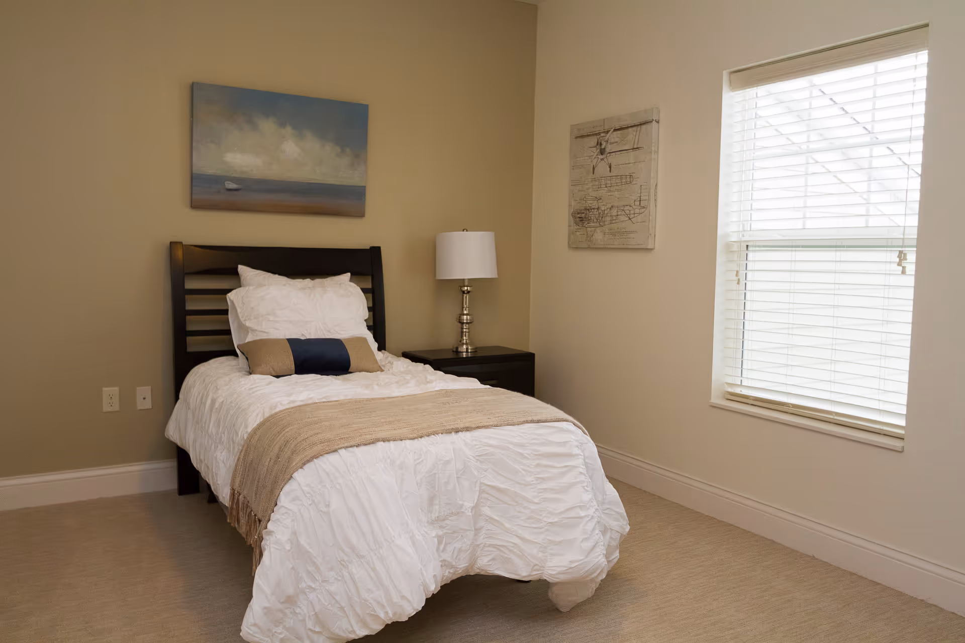 A small bedroom with a single bed made with white bedding and a beige throw blanket. There is a dark wooden headboard, a nightstand with a silver lamp, a window with white blinds, and two pieces of wall art.