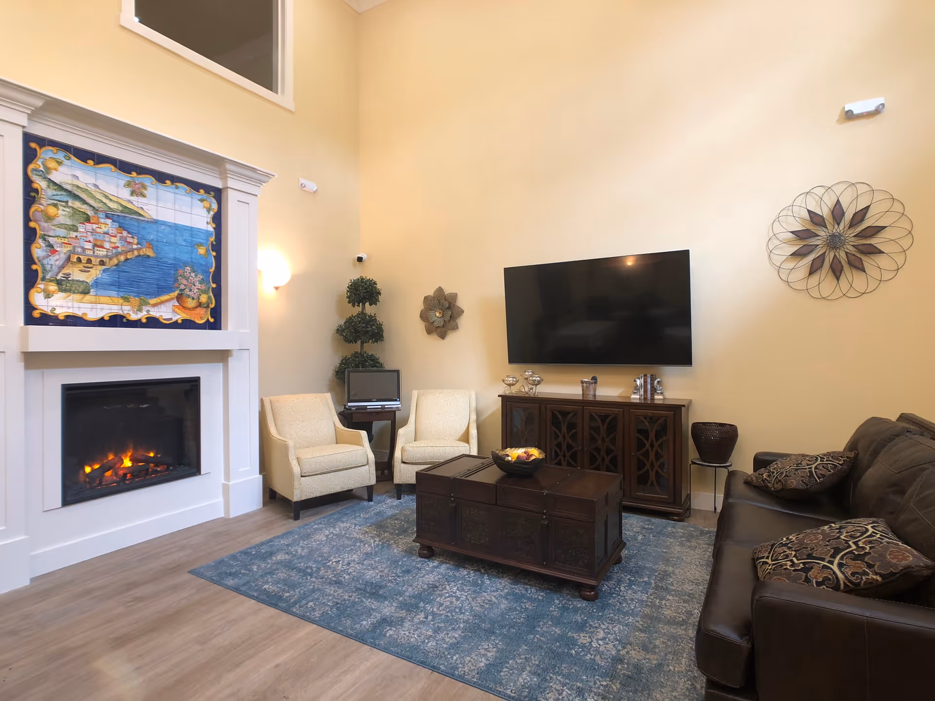 A cozy living room with a lit fireplace featuring a colorful tile mural above it, two beige armchairs next to a small table with a computer, a large flat-screen TV mounted on the wall above a wooden cabinet, a dark brown leather sofa with patterned cushions, a decorative wall piece, and a blue area rug on a wooden floor.