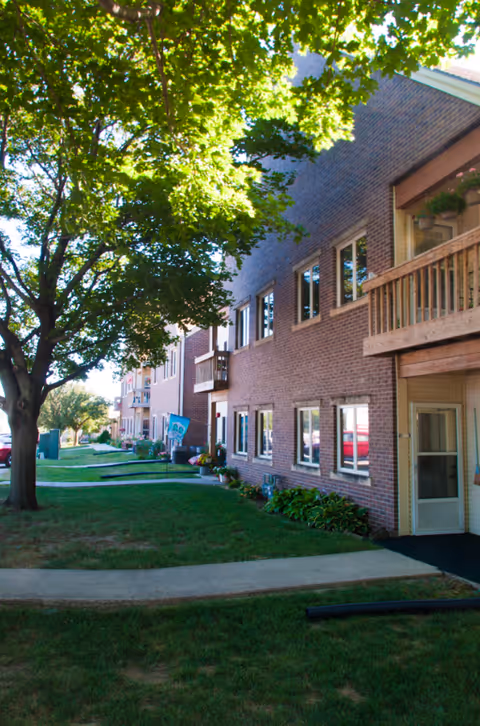 Exterior view of a multi-story brick senior living community building with balconies, windows, and a green lawn with a large tree in the foreground. A sidewalk runs along the building with some plants near the entrance.