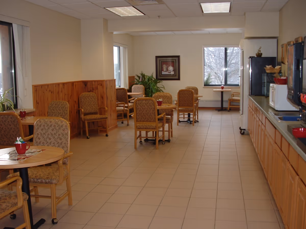 A dining area in an assisted living facility with several round tables and cushioned chairs arranged neatly. The room has large windows letting in natural light, a tiled floor, and a kitchenette area with a microwave, refrigerator, and cabinets along one wall. There are plants and framed artwork on the walls.