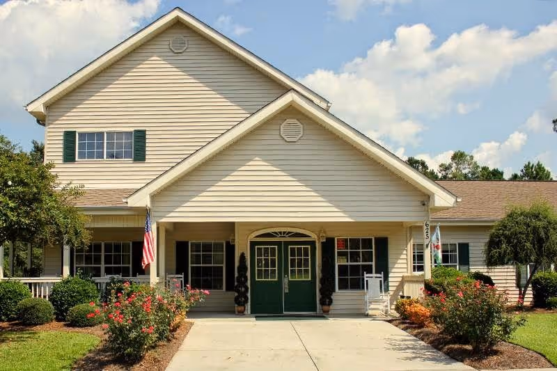 Front exterior view of a two-story senior living facility building with beige siding, green double doors, green window shutters, an American flag, and well-maintained landscaping including bushes and flowering plants.