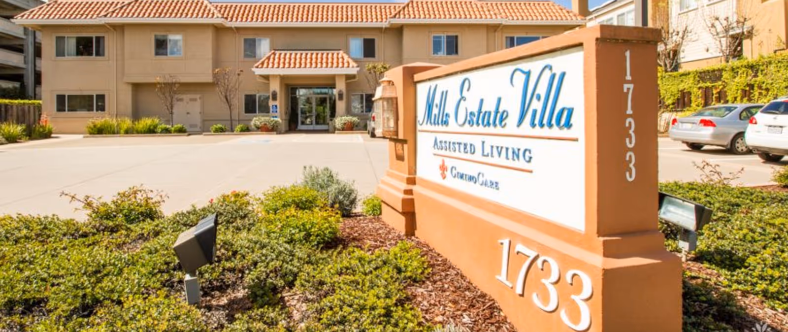 Entrance and sign for Mills Estate Villa assisted living with the building, driveway, and landscaped grounds visible.