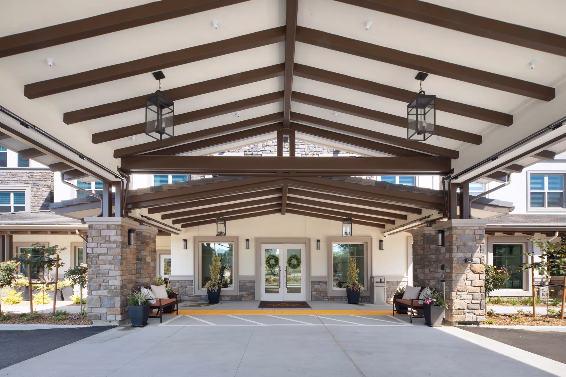 Covered entrance of a senior living facility with stone pillars, wooden beams on the ceiling, hanging lantern-style lights, benches with cushions, potted plants, and glass double doors decorated with wreaths.