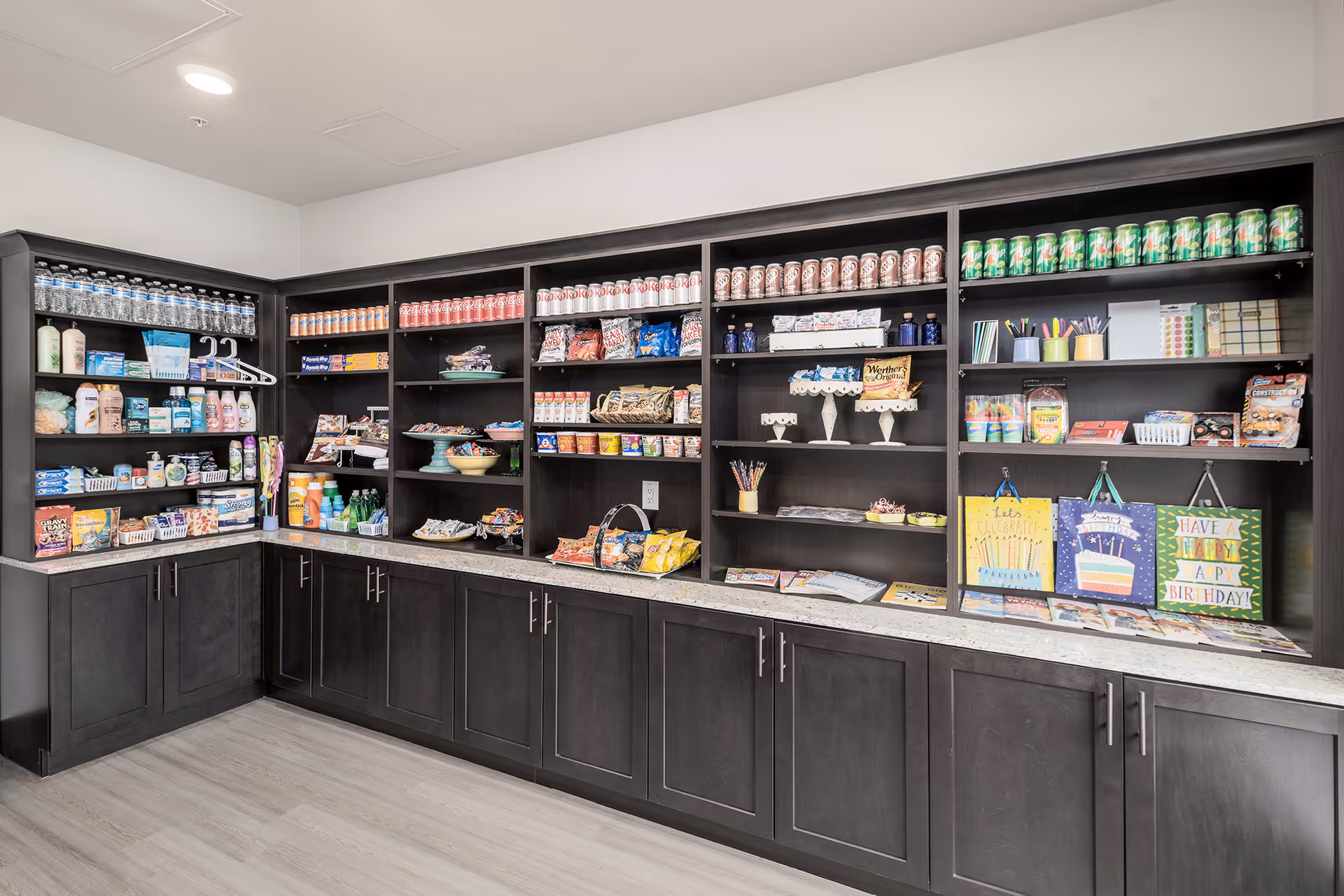 A well-organized pantry or snack area with dark wooden shelves and cabinets. The shelves are stocked with bottled water, soda cans, snacks, toiletries, gift bags, and craft supplies. The countertop is clear, and the floor has light-colored wood flooring.