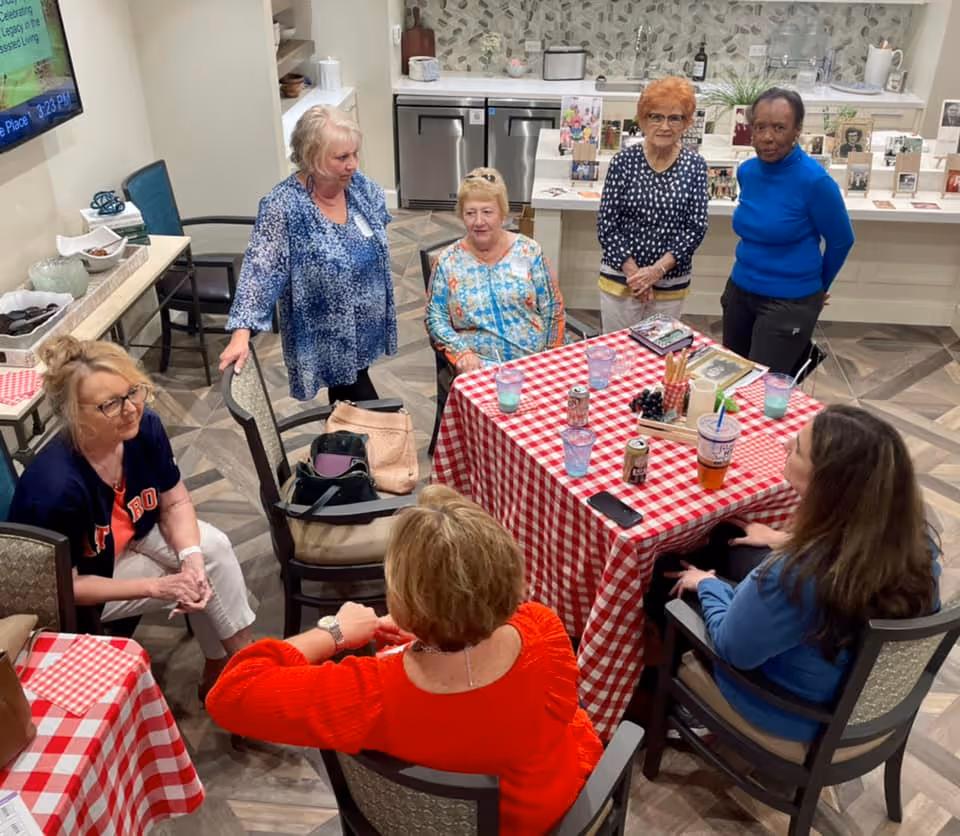 Several older women gathered around a red-and-white checkered table in a senior living community common room.