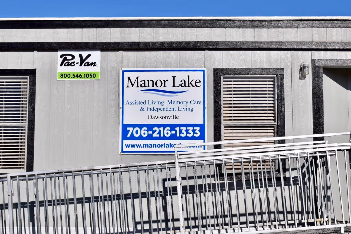 Exterior wall of a building with two windows and a door, featuring a sign for Manor Lake Assisted Living, Memory Care & Independent Living in Dawsonville, along with a phone number and website. A metal ramp is visible in front of the building.