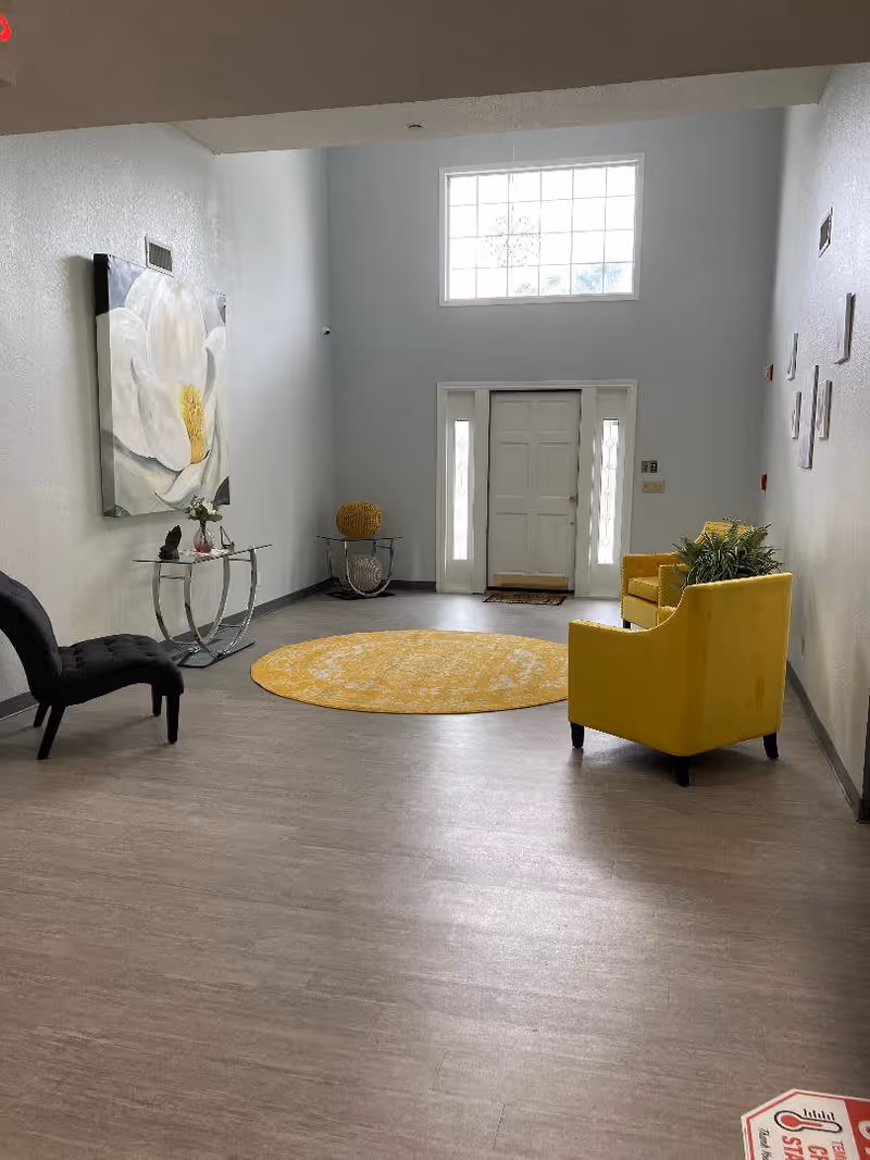 A spacious interior room with a high ceiling and a large window above a white front door. The room features a round yellow rug in the center, two yellow armchairs with a plant on one, a black cushioned chair, a glass console table with decorative items, and a large floral painting on the wall. The floor is light wood, and there are several small framed pictures on the right wall.