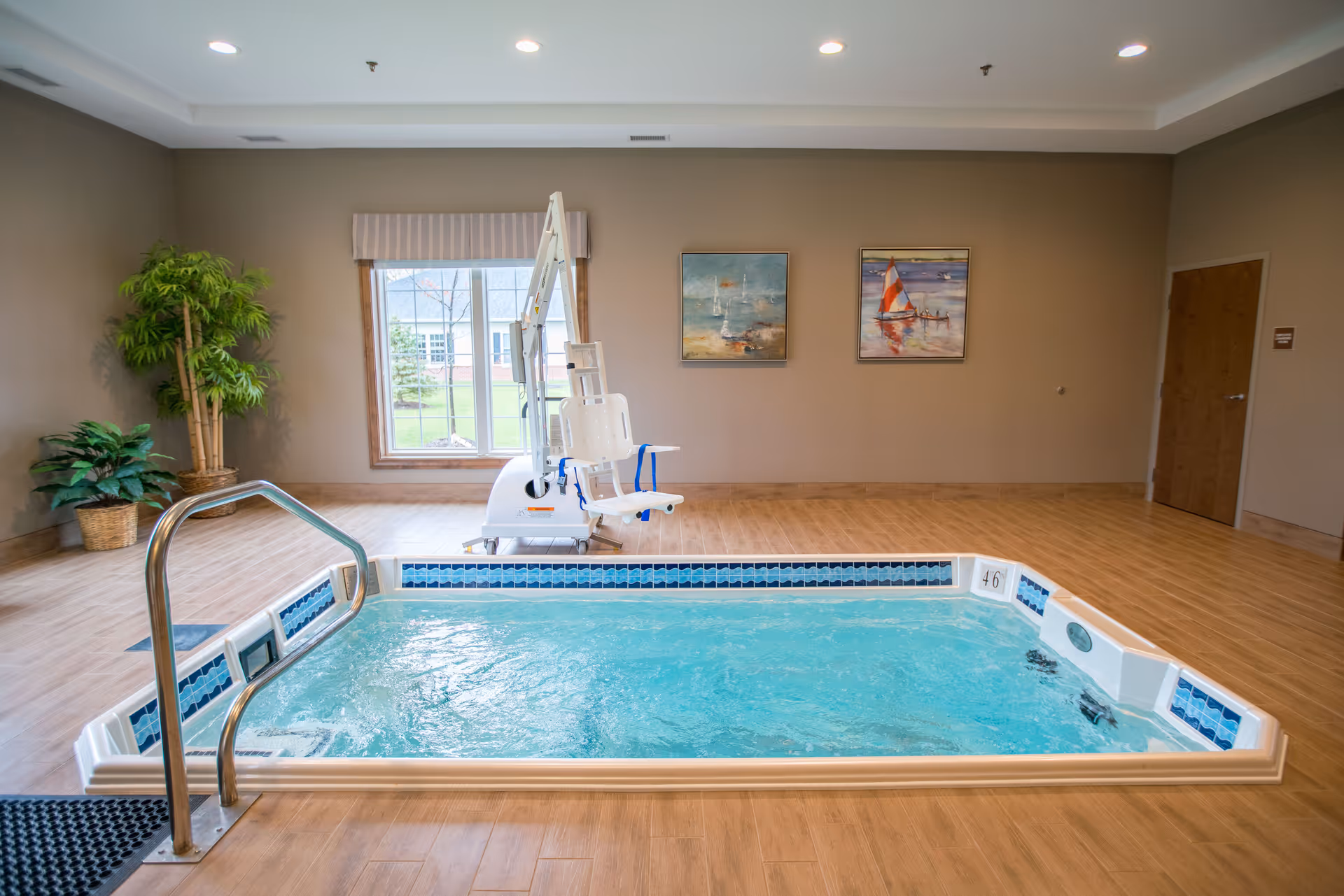 Indoor therapy pool with bubbling water, a metal handrail, and a white pool lift chair. The room has wooden flooring, beige walls, two framed paintings of sailboats, a window with a view of greenery outside, and two potted plants in the corner.