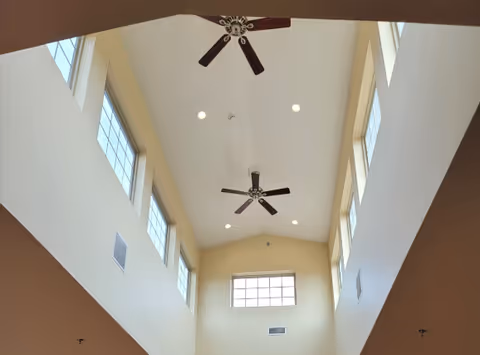 View looking up at a high ceiling with multiple windows on both sides, two ceiling fans, and recessed lighting in a bright interior space.