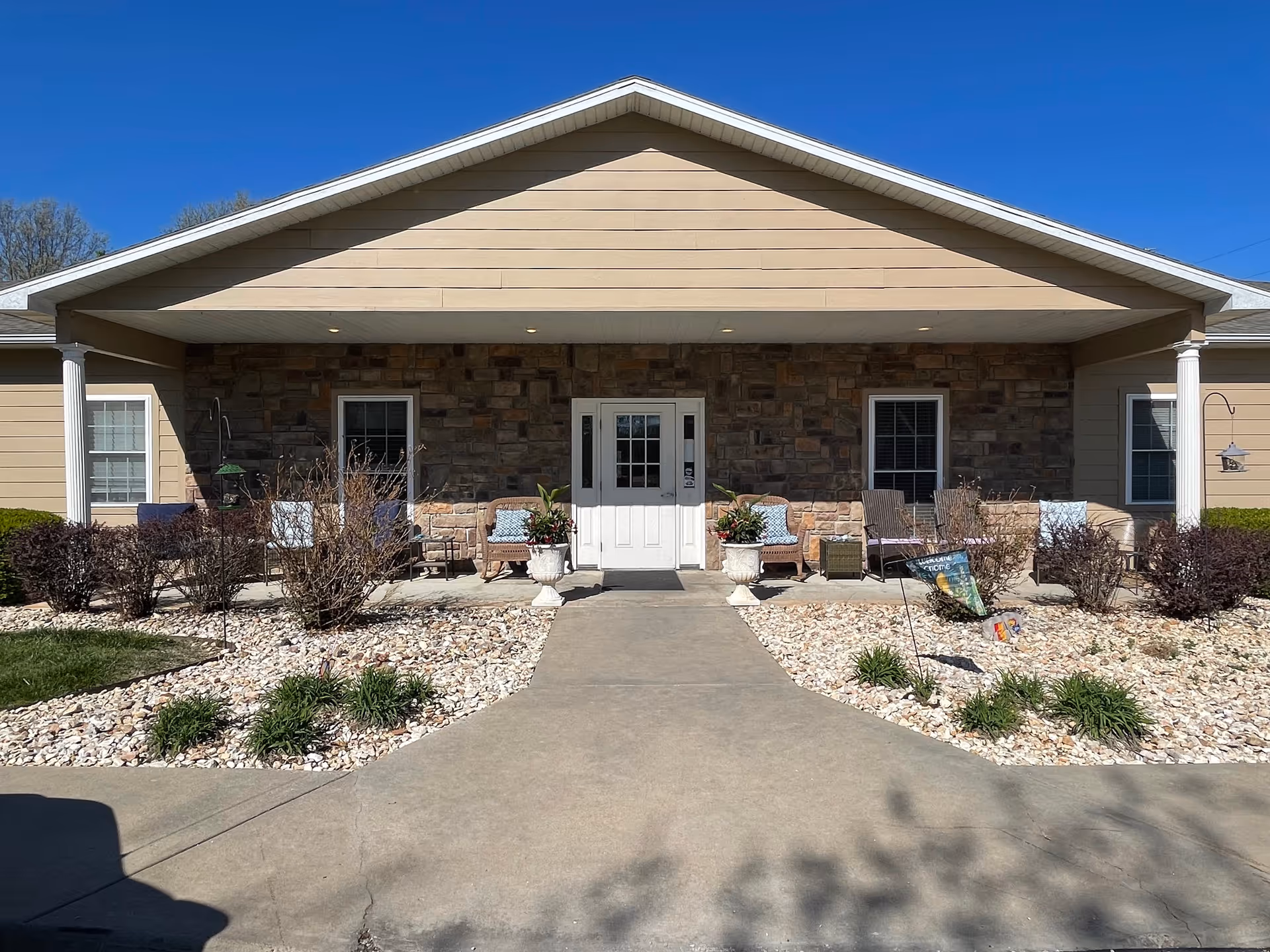 Front entrance of Oakview Estates featuring a covered porch with stone and beige siding, white double doors, outdoor seating with chairs and cushions, potted plants, and landscaping with rocks and shrubs.