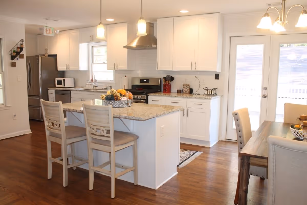 Bright and modern kitchen with white cabinets, stainless steel appliances including a refrigerator, microwave, and stove. A kitchen island with two chairs and a decorative centerpiece is in the foreground. To the right, a dining table with upholstered chairs is partially visible near glass doors leading outside.