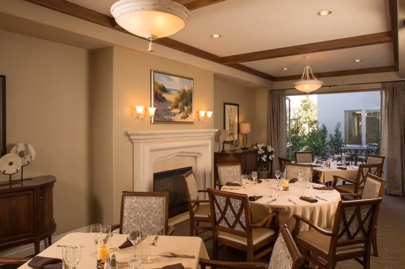 A warmly lit dining room with round tables covered in beige tablecloths, set with glassware, napkins, and cutlery. The room features a white fireplace with a painting above it, wooden chairs with patterned upholstery, and a sideboard with decorative items. Large windows with curtains reveal an outdoor patio area with additional seating.