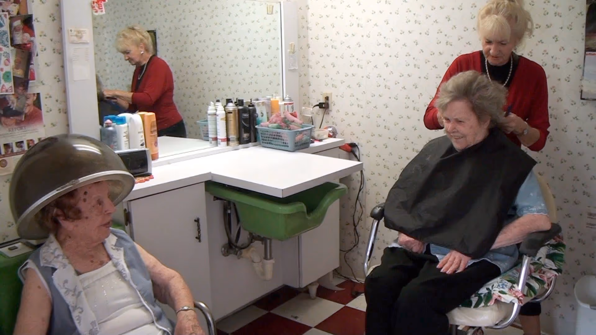 Two elderly women in a hair salon setting; one woman is sitting under a hair dryer while the other is having her hair styled by a hairdresser. The room has floral wallpaper, a large mirror, and various hair care products on the counter.