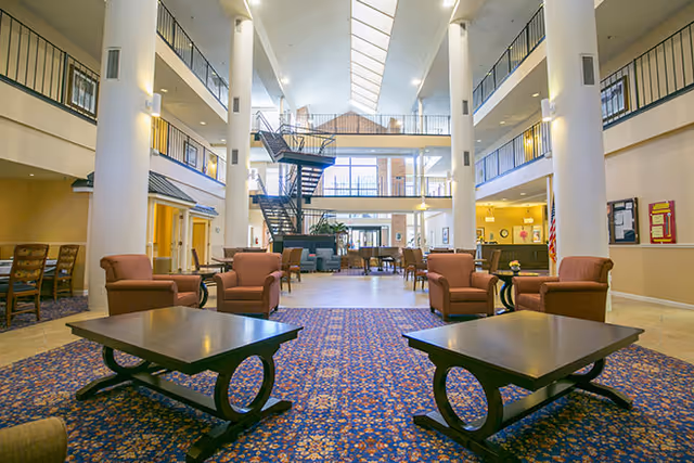 Spacious and well-lit common area in a senior living facility with multiple seating arrangements including armchairs and coffee tables on a patterned carpet. The room features high ceilings with skylights, large white columns, a staircase leading to an upper level, and a reception desk in the background.