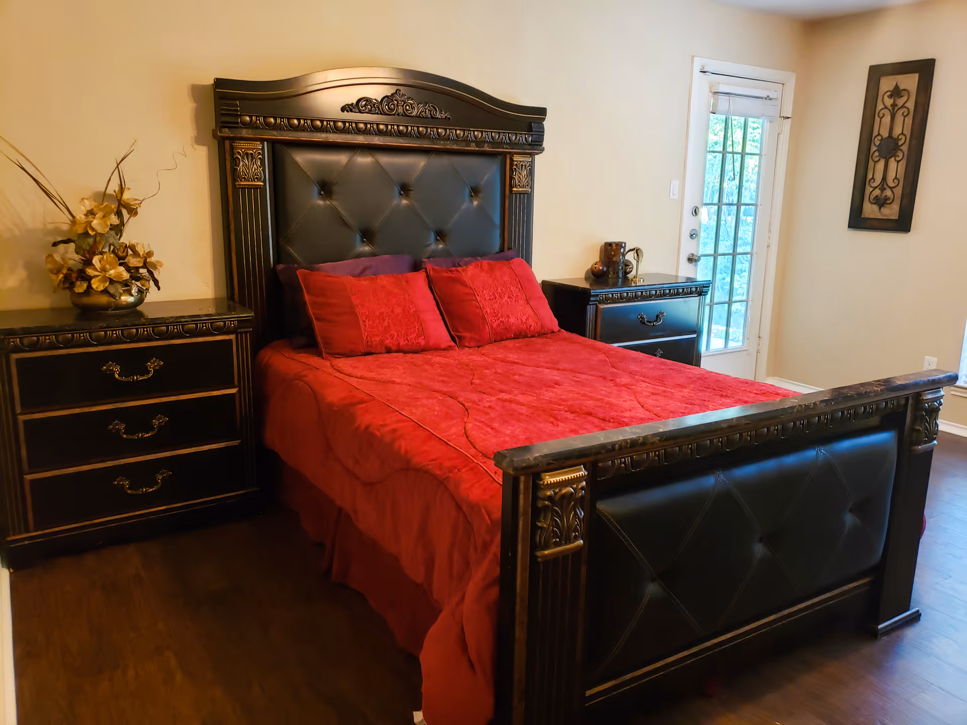 Ornate dark wood bed with a tufted black headboard and red bedding flanked by matching nightstands in a bedroom with a glass door.