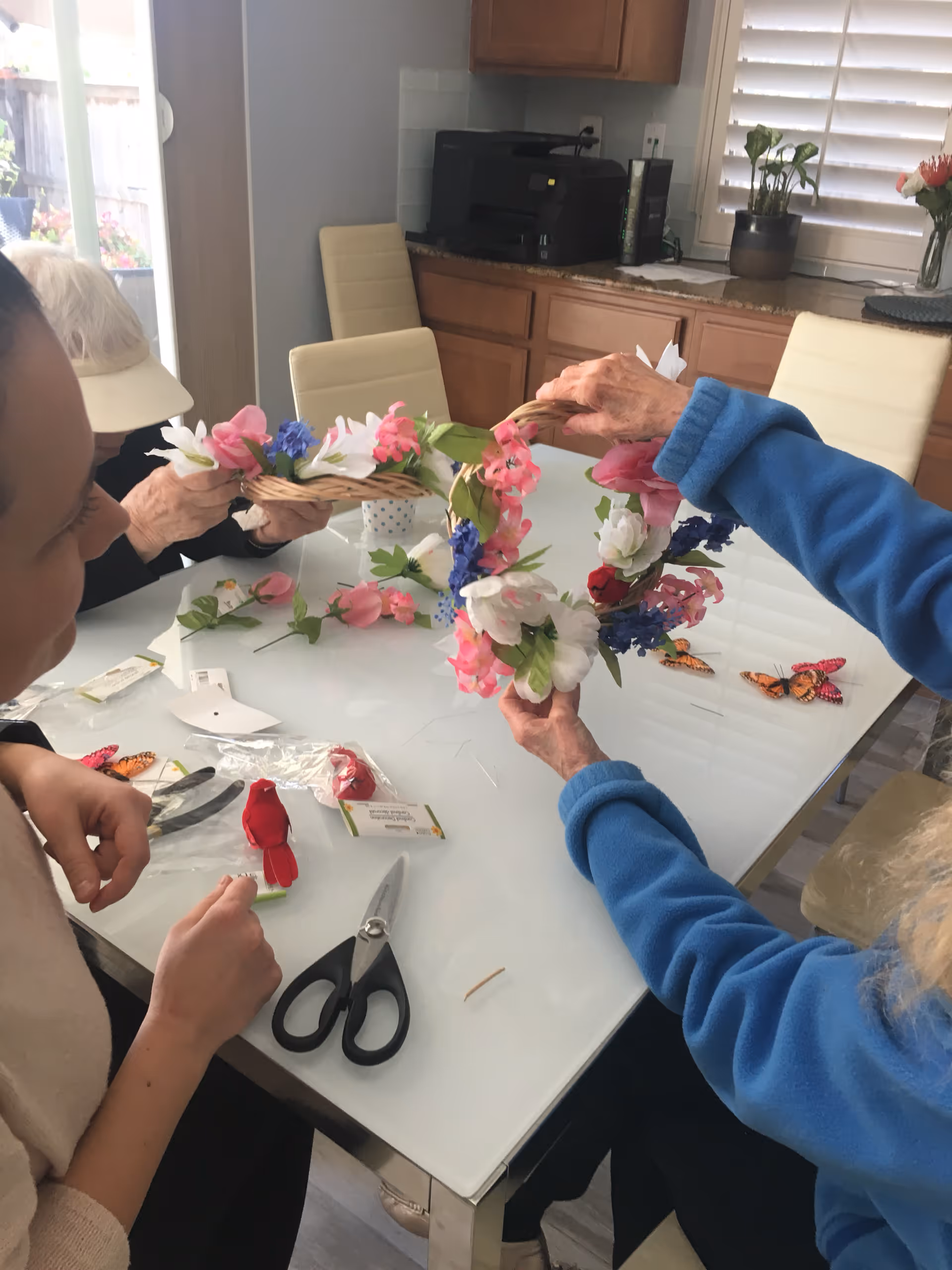 Several people seated around a glass-topped table crafting a floral wreath in a kitchen/dining area.