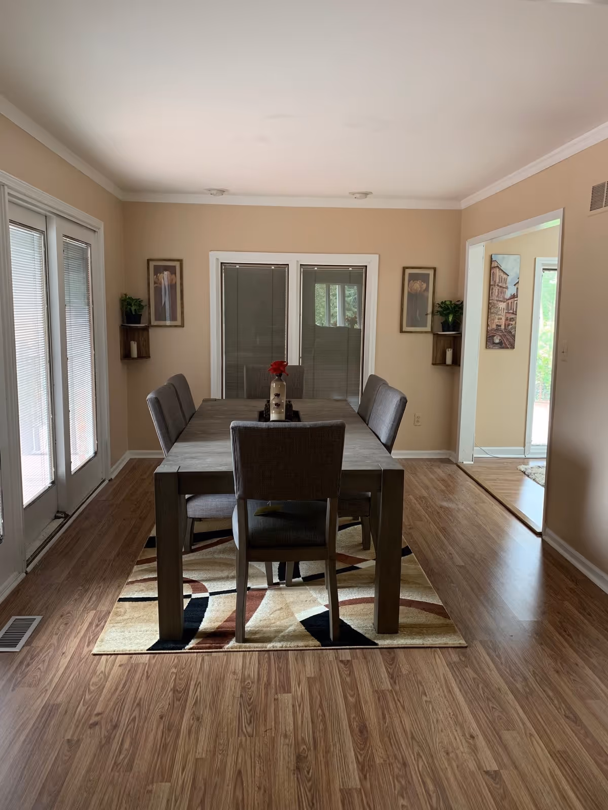 A dining room with a rectangular wooden table and six gray upholstered chairs arranged around it. The table is placed on a patterned area rug with beige, black, and brown colors. There are two framed pictures and small shelves with plants and candles on the beige walls. Double glass doors with blinds are on the left side, and another set of glass doors with blinds is straight ahead. The floor is wood laminate, and the room has a clean, simple, and cozy appearance.
