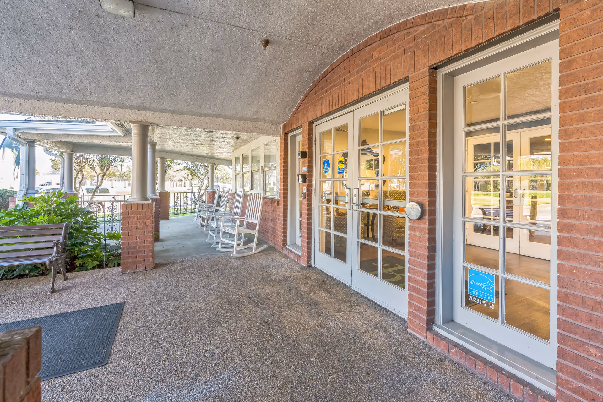 Covered outdoor patio area with brick pillars and white rocking chairs lined up along the wall. There is a wooden bench on the left side near green plants. Glass double doors with white frames lead inside the building, showing a glimpse of an interior room with chairs and tables.