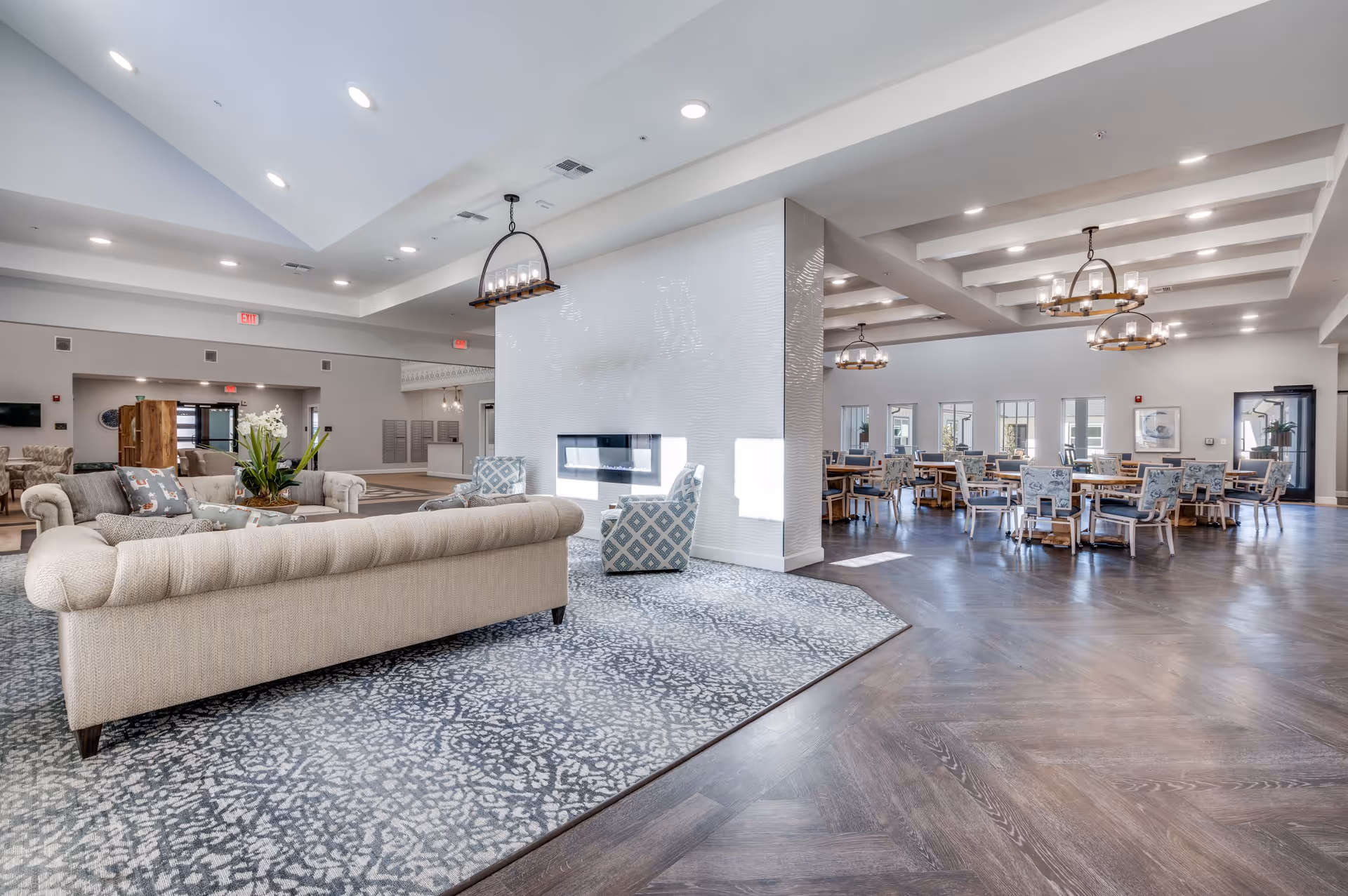 A spacious and well-lit common area in a senior living facility featuring a cozy seating area with a beige sofa, patterned armchairs, a modern fireplace, and a dining area with multiple tables and chairs. The room has high ceilings with recessed lighting and decorative chandeliers, wood flooring, and large windows allowing natural light.