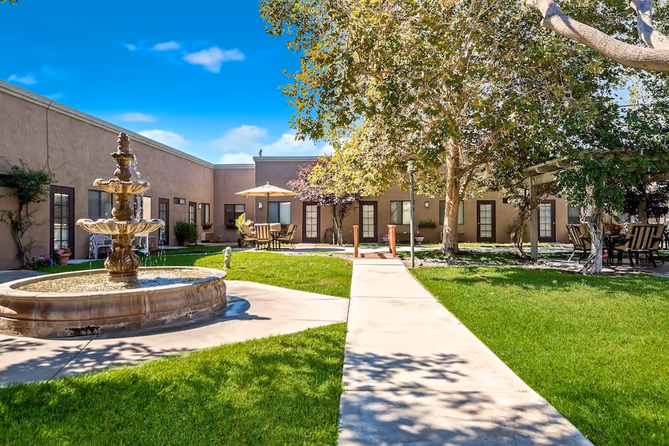 Outdoor courtyard area at Sierra Vista Independent & Assisted Living featuring a multi-tiered stone fountain, green grass, a concrete walkway, patio tables with umbrellas, and trees providing shade.