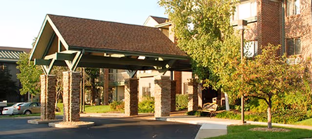 Covered entrance area with stone pillars and a peaked roof at the front of a senior living facility building, surrounded by trees and greenery with a paved driveway and parked cars nearby.