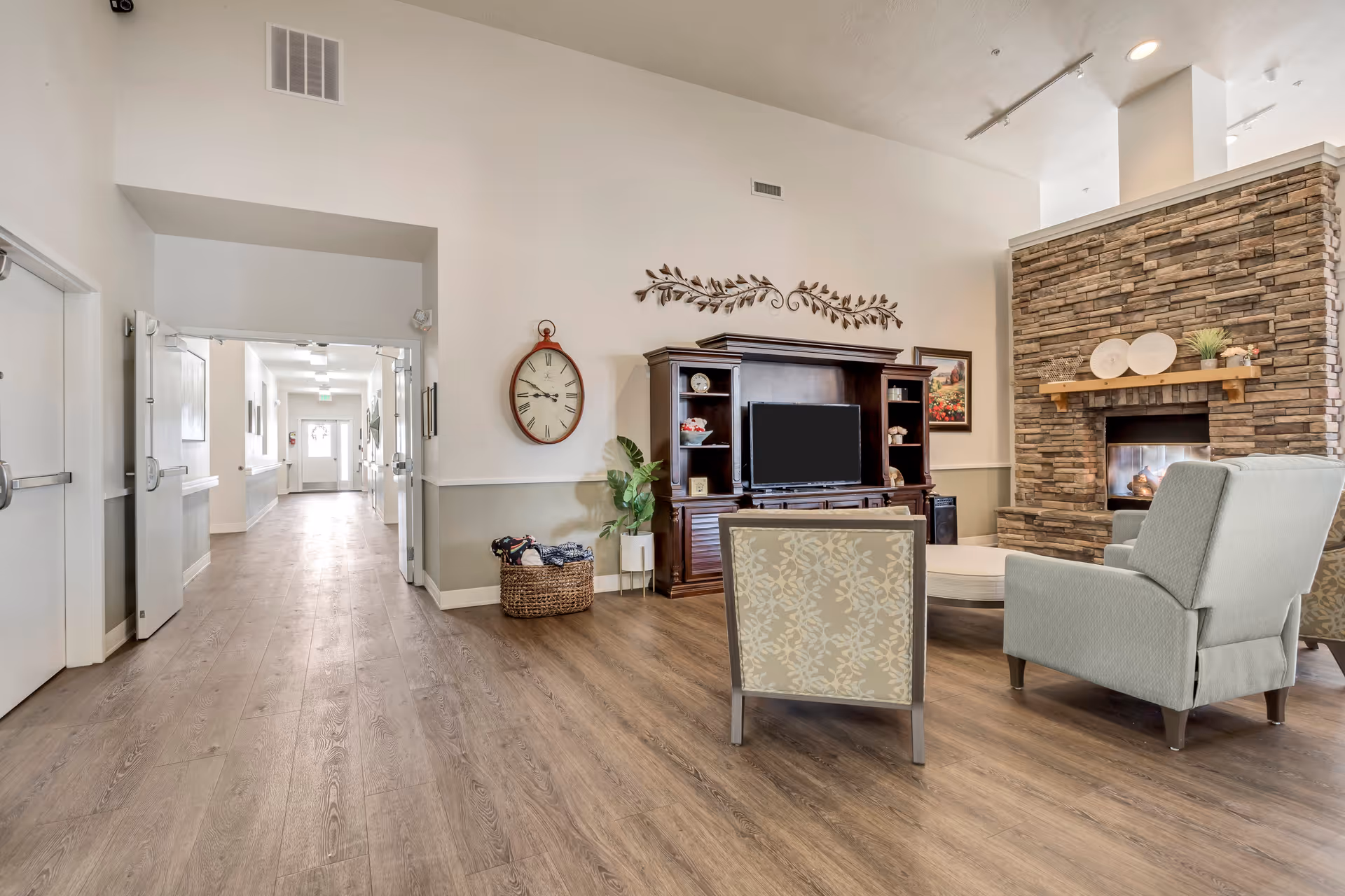 A spacious and well-lit living room area in a senior living facility with wooden flooring, a stone fireplace, a large wooden entertainment center with a TV, two upholstered chairs, and decorative wall art. A hallway with open double doors leads to other parts of the facility.
