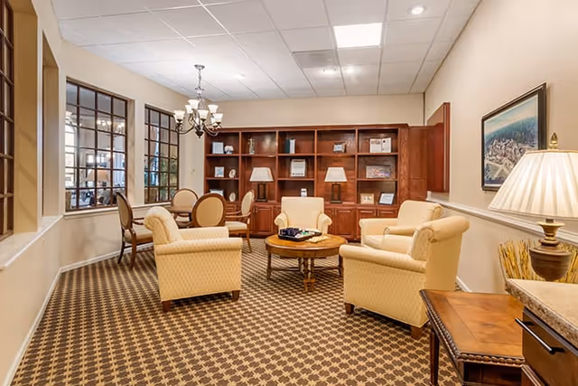 A cozy living room area with four beige upholstered armchairs arranged around a round wooden coffee table. Behind the seating area is a large wooden bookshelf with various decorative items and two table lamps. To the left, there is a dining area with a round table and chairs visible through large interior windows. The room has patterned carpet flooring, beige walls, and a ceiling with recessed lighting and a chandelier.