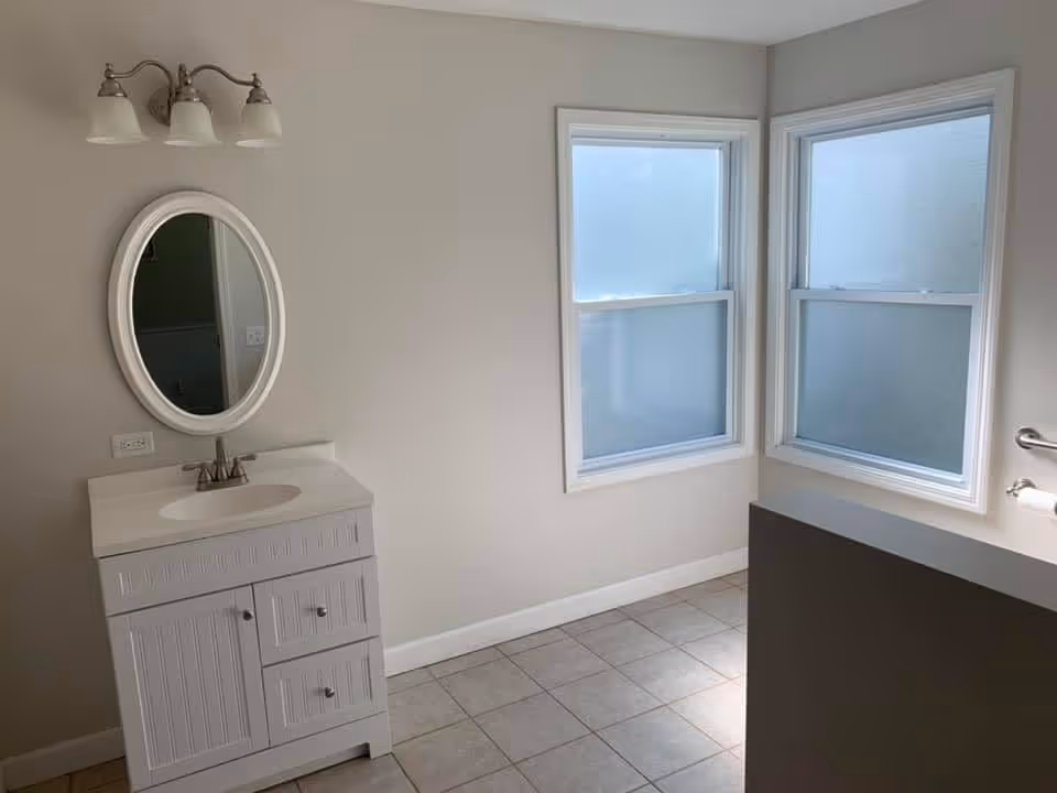A bathroom with a white vanity cabinet featuring a sink and an oval mirror above it. There are two frosted windows on the adjacent wall allowing natural light to enter. The floor is tiled, and a toilet paper holder is mounted on the right wall.