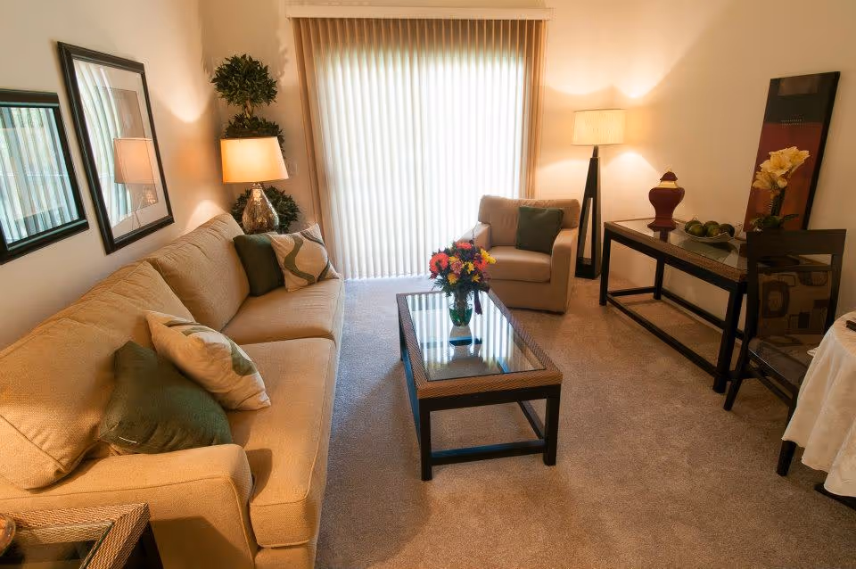 Cozy living room with a beige sofa, armchair, glass-top coffee table, lamps and vertical blinds covering a sliding door.