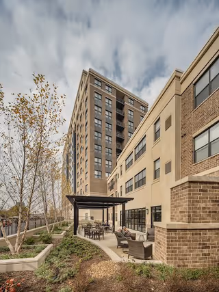 Outdoor patio area at Clarendale Clayton with seating arrangements including chairs and tables under a pergola, adjacent to a multi-story brick and beige building, with landscaped garden beds and young trees under a cloudy sky.
