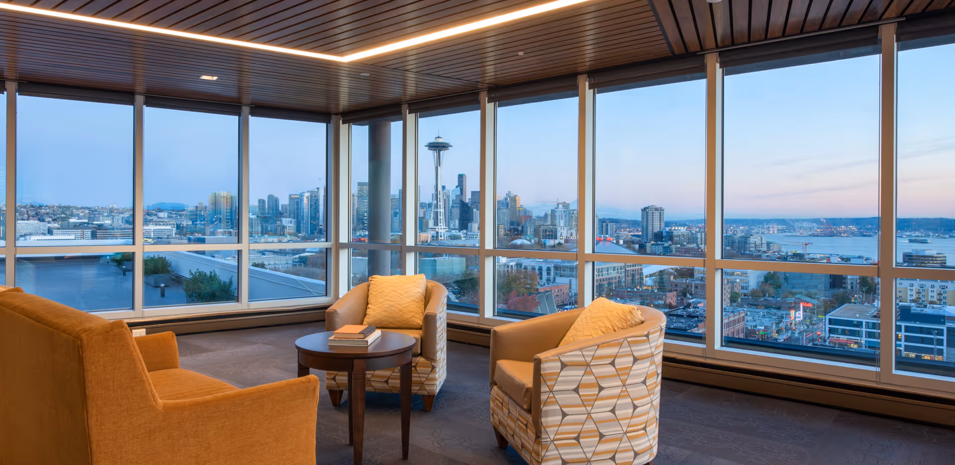 A cozy seating area inside Bayview Retirement Community with two patterned armchairs and one orange sofa arranged around a small round wooden table with books on it. Large floor-to-ceiling windows provide a panoramic view of the Seattle skyline, including the Space Needle, with a clear sky at dusk.