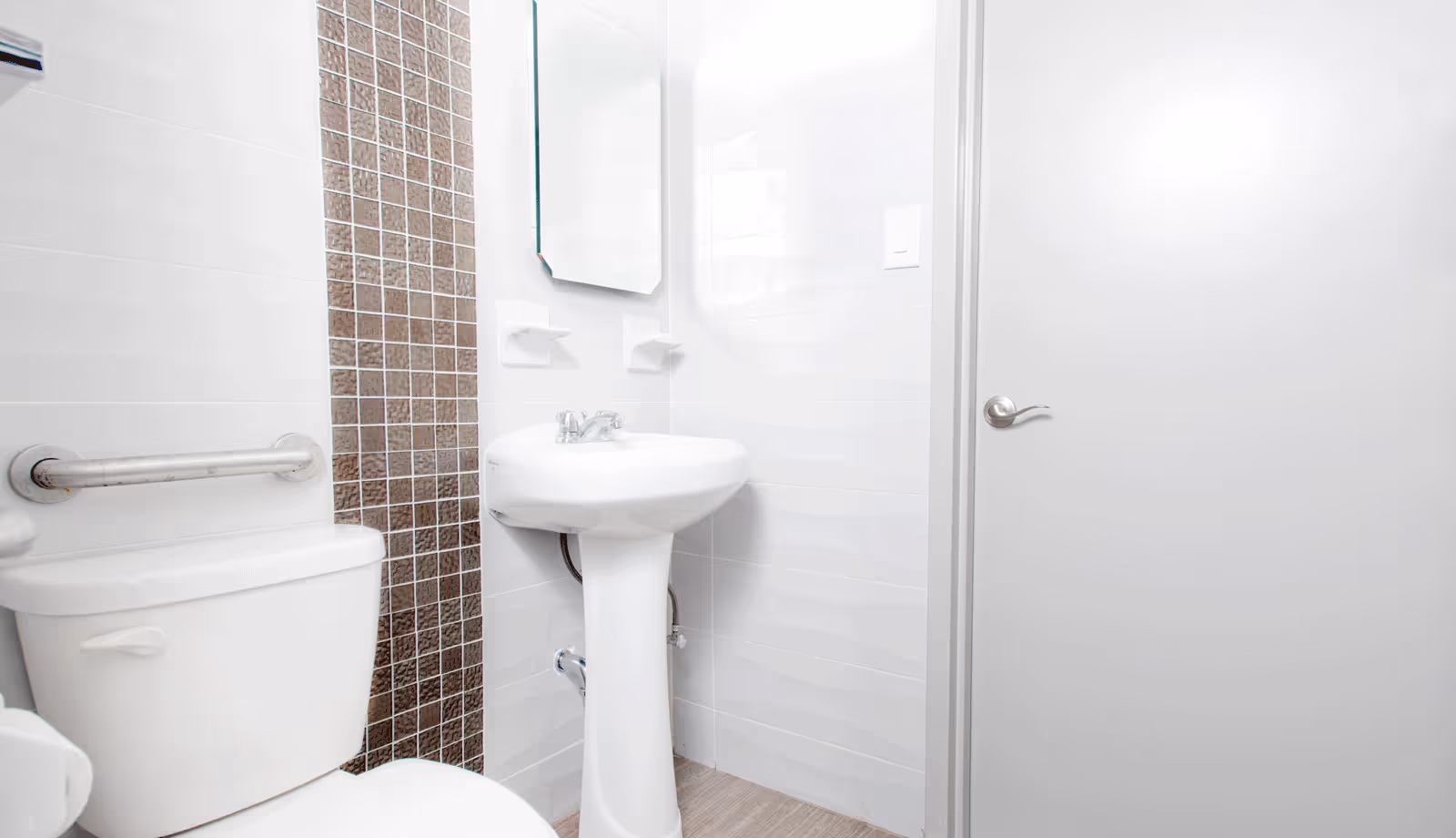 A clean and bright bathroom featuring a white toilet with a metal grab bar, a white pedestal sink with a faucet, a rectangular mirror above the sink, and a door with a silver handle. The walls are tiled in white with a vertical strip of small brown mosaic tiles behind the sink.