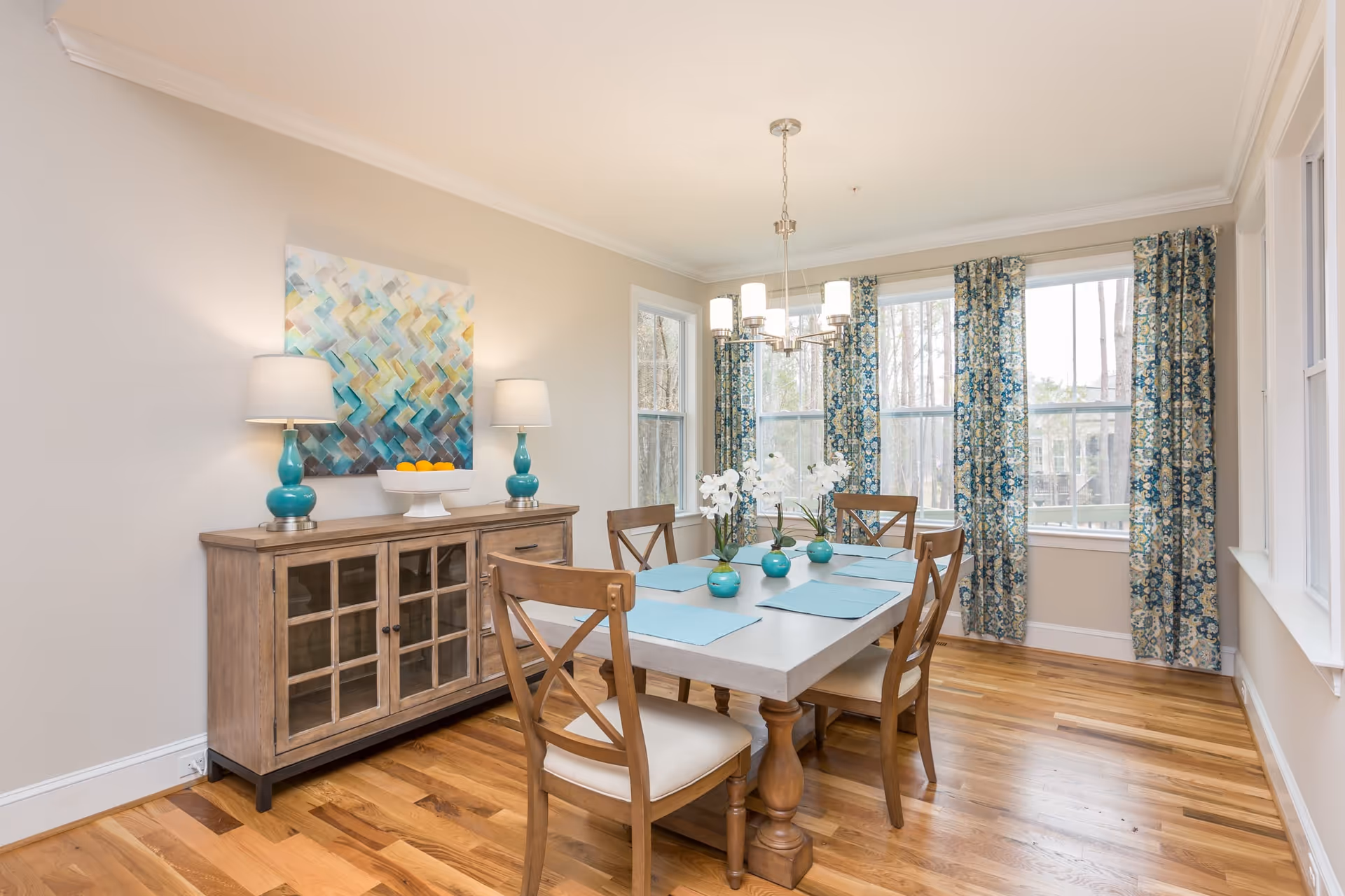 Bright dining room with a wooden table and chairs, teal accents and vases, a buffet with lamps, and large windows with patterned curtains.