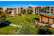 Exterior view of a multi-story senior living facility named The Community at Rockhill, featuring a large green lawn, landscaped gardens, walking paths, and a covered outdoor pavilion under a clear blue sky.