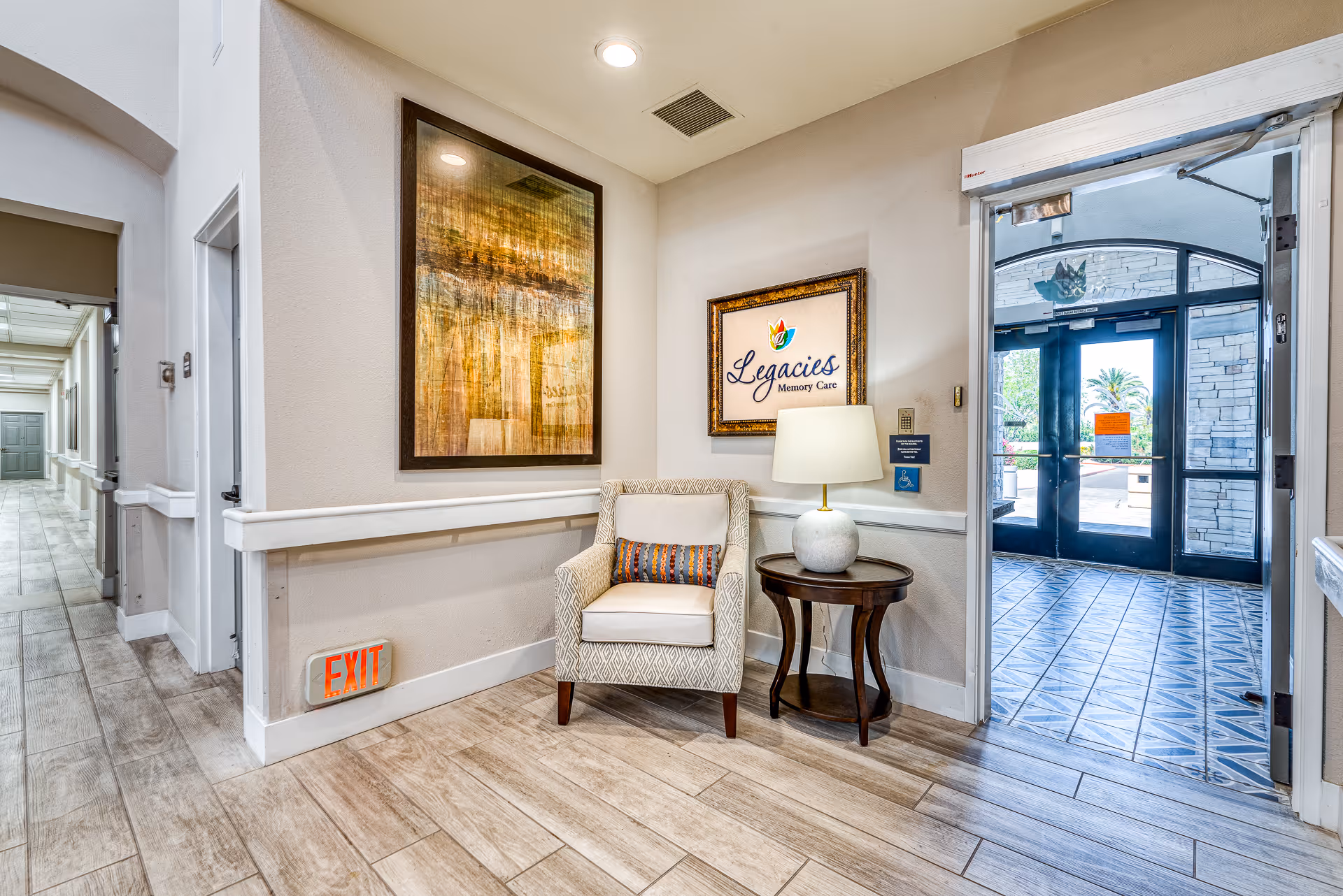 Small reception seating area with an upholstered armchair, side table and lamp beside a framed 'Legacies Memory Care' sign and an open doorway to the exterior.