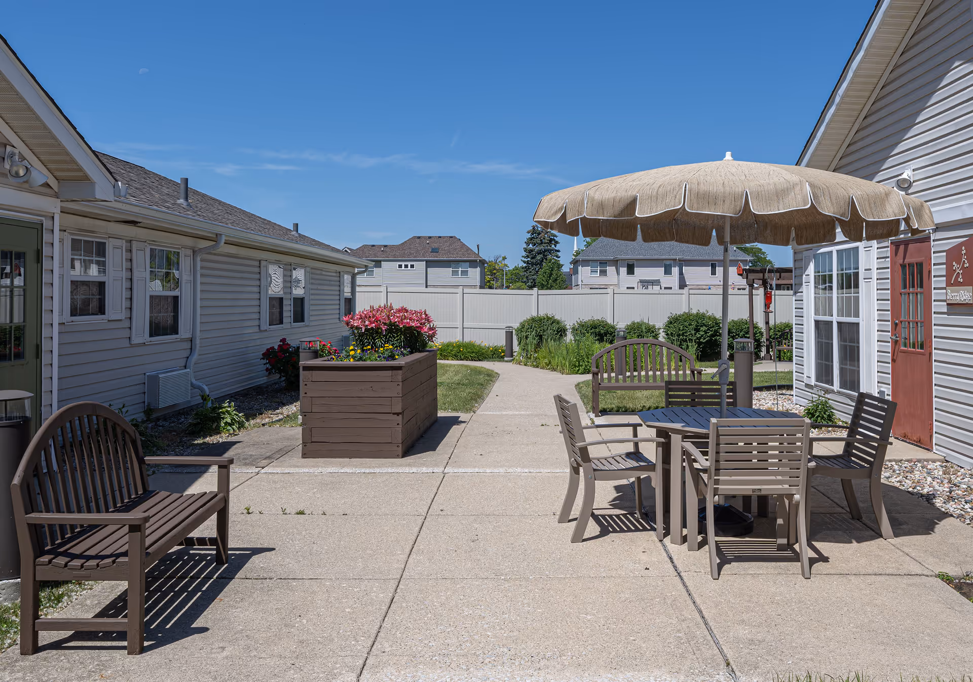Outdoor patio area with brown benches and a table with four chairs under a large beige umbrella. There are flower planters and shrubs along the pathway, with residential buildings and a clear blue sky in the background.