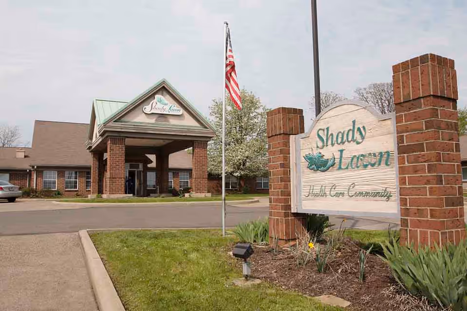 Exterior view of The Oaks at Shady Lawn health care community building with a brick entrance, a covered driveway, an American flag on a flagpole, and a wooden sign with the community name surrounded by landscaping.