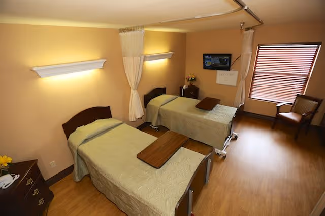 A shared bedroom in a healthcare facility with two single beds, each with a beige bedspread and a wooden overbed table. The room has warm beige walls, wooden flooring, a window with closed blinds, a wooden chair, a small nightstand with flowers, and a wall-mounted TV.