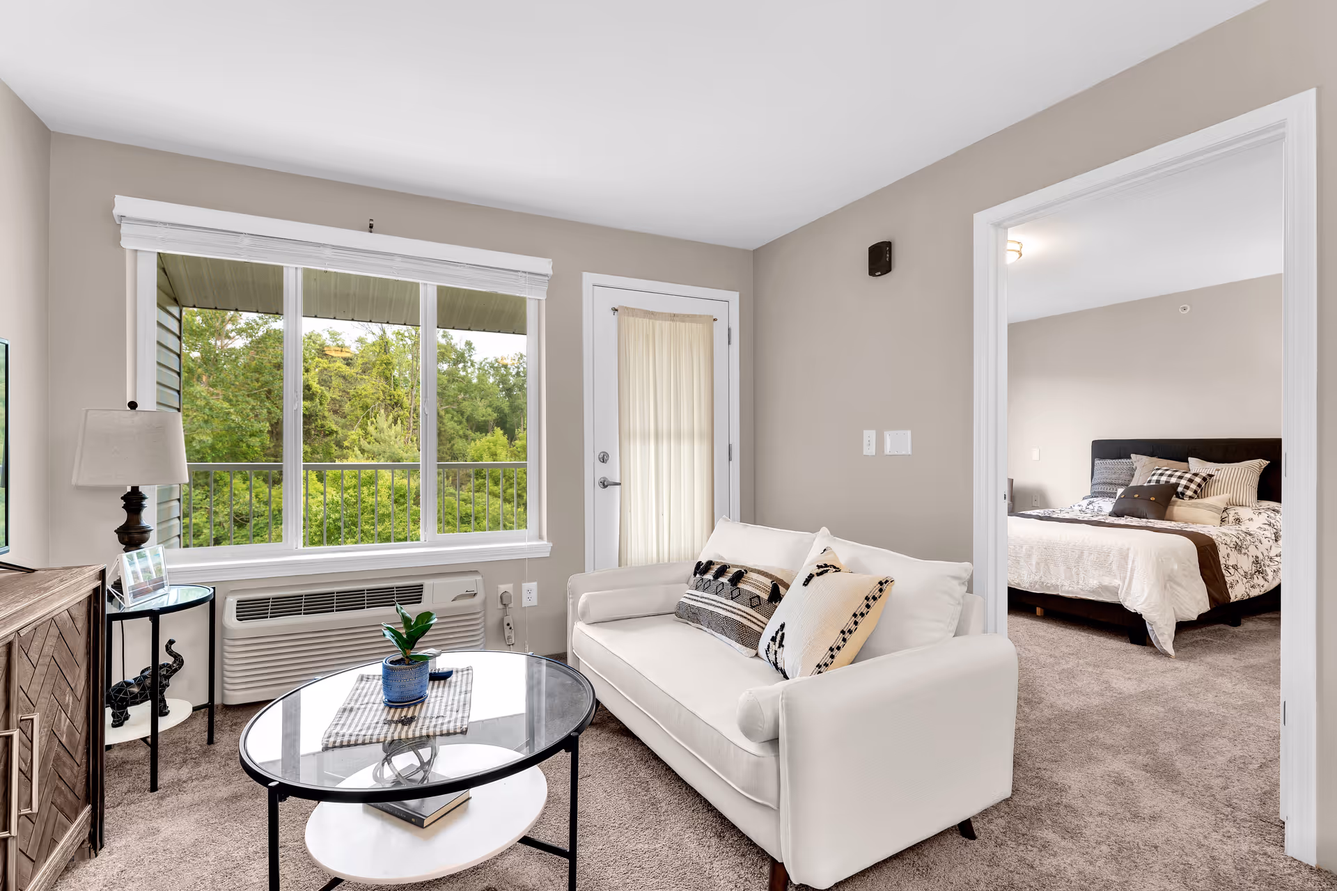 A cozy living room with a white couch adorned with patterned pillows, a round glass coffee table with a small plant, a side table with a lamp and decorative elephant figurines, and a large window showing green trees outside. A door with a sheer curtain leads to a balcony, and an open doorway reveals a bedroom with a bed covered in a white and brown patterned comforter.