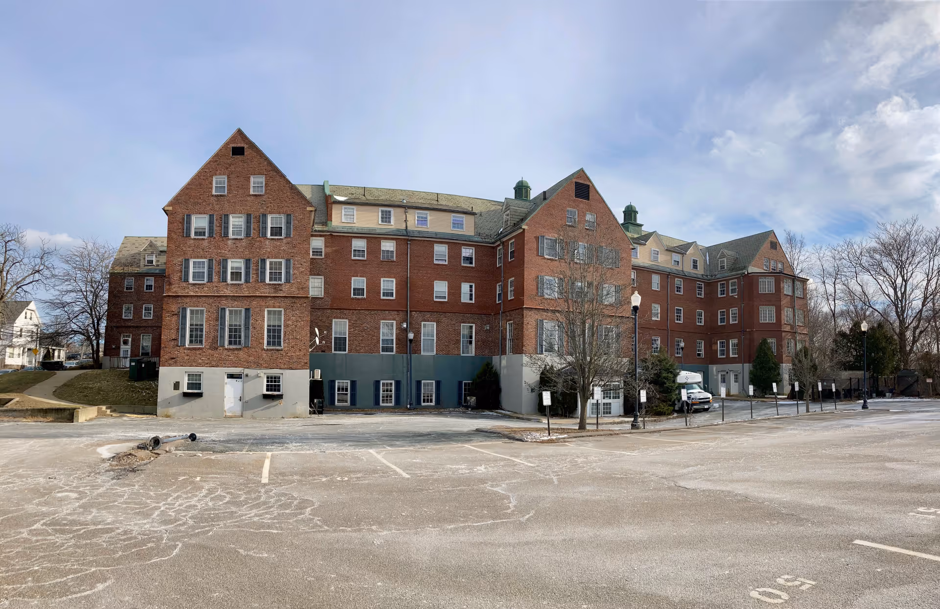 Exterior view of a large multi-story brick building with multiple windows and a greenish roof, surrounded by a mostly empty parking lot with some trees and street lamps under a partly cloudy sky.