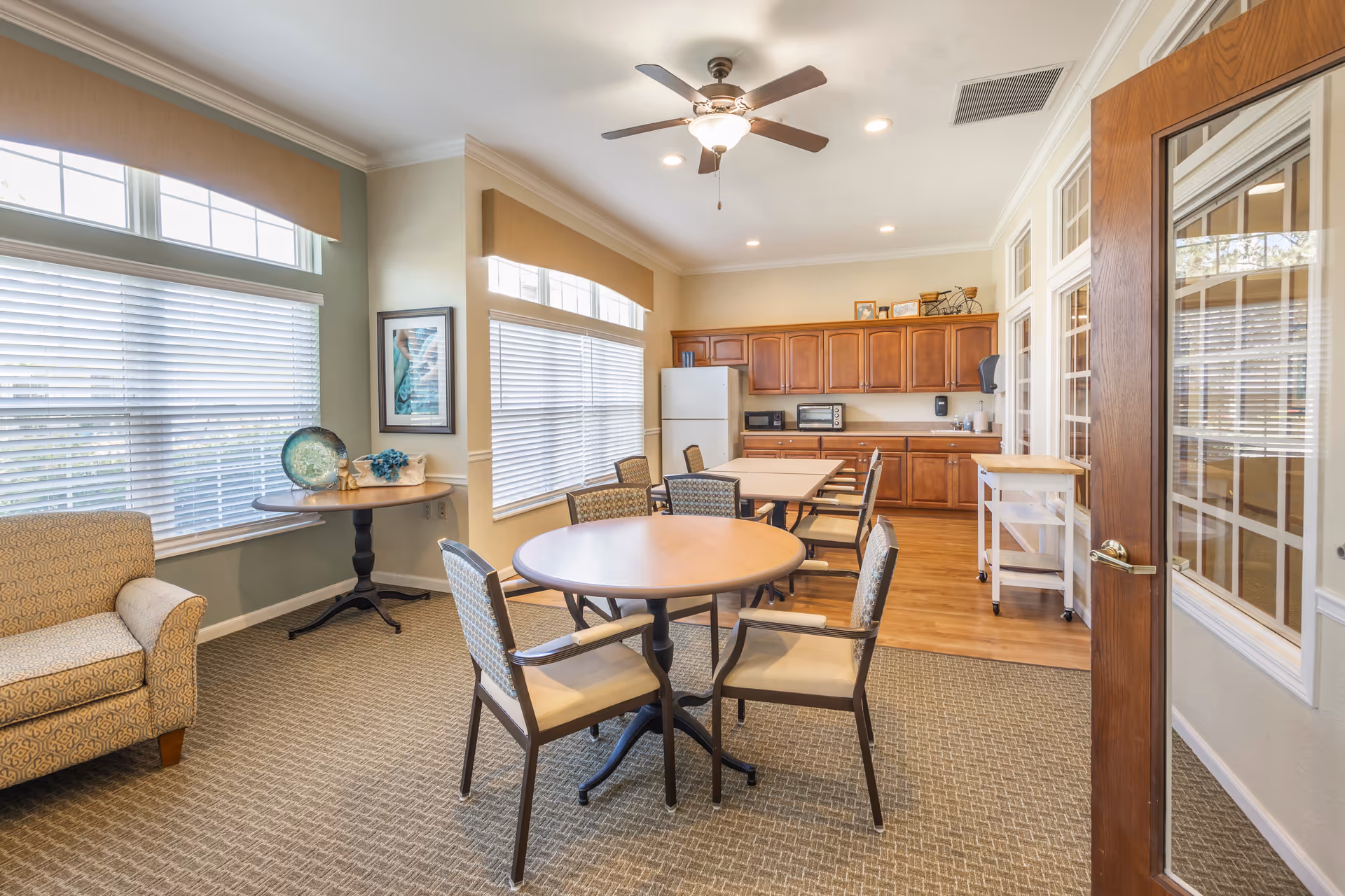 A bright and spacious common area in a senior living facility featuring a round table with four cushioned chairs, a small round side table with decorative items, a comfortable armchair, and a kitchenette with wooden cabinets, a refrigerator, a toaster oven, and a microwave. Large windows with blinds allow natural light to fill the room, and a ceiling fan with a light fixture is mounted on the ceiling.