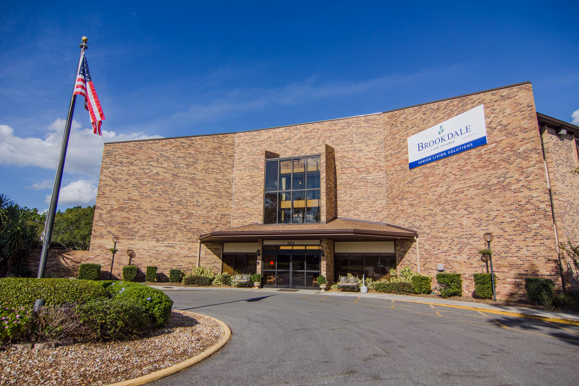 Exterior front view of a brick building with a large entrance and a sign that reads 'Brookdale Lake Tavares Senior Living Solutions'. An American flag is flying on a flagpole to the left, and there are bushes and landscaped areas around the driveway leading to the entrance.