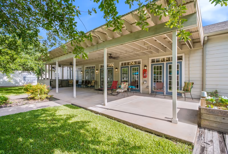 Covered exterior patio with chairs and tables in front of multiple French doors at a senior living facility.