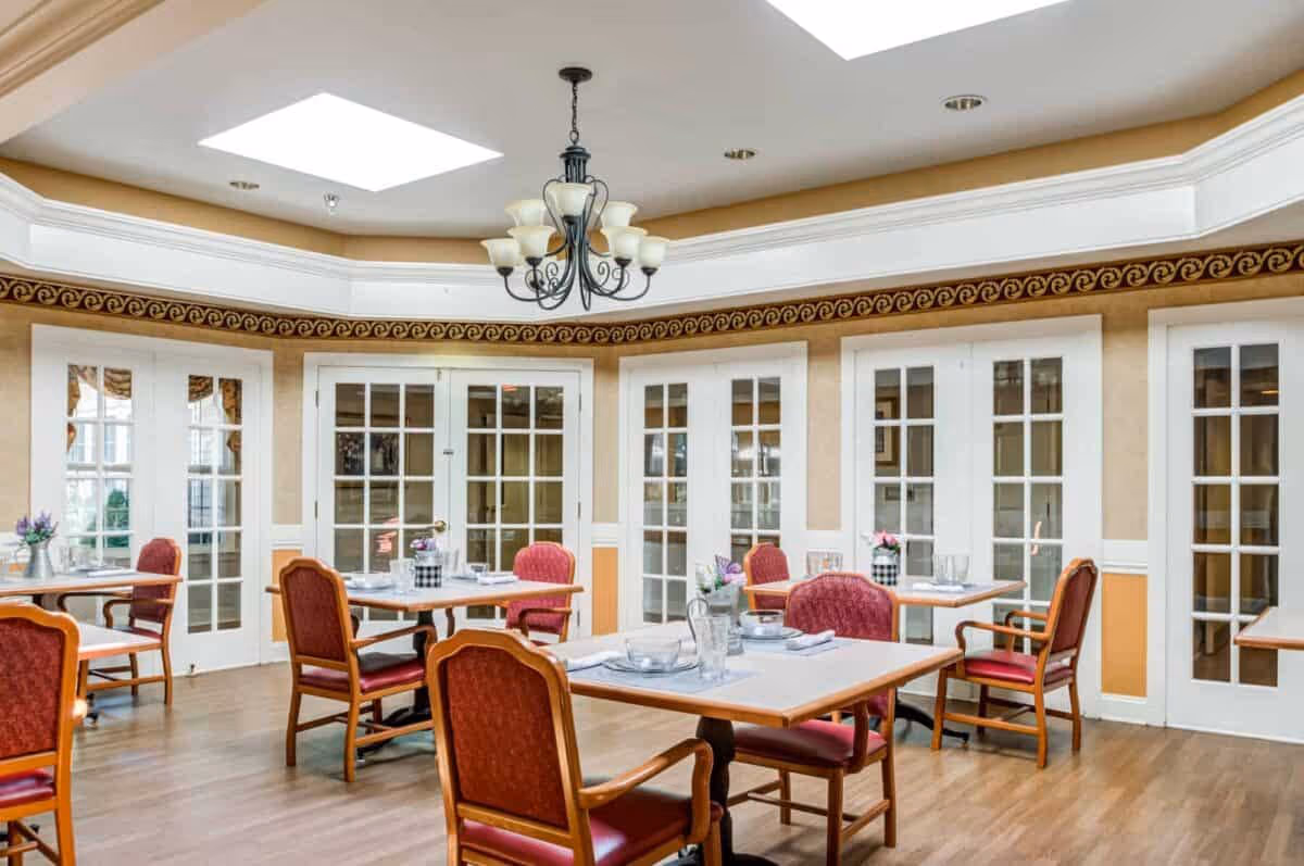 Bright dining room with multiple tables and red upholstered chairs, a central chandelier, and a wall of French doors.